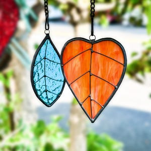 Pode incluir: Dois ornamentos de folhas de vitral. Um tem a forma de folha em tons de laranja, o outro tem a forma de lágrima em tons de azul. Ambos têm bordas de metal preto e são suspensos por correntes pretas.