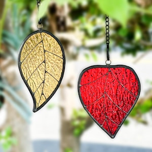 May include: Two stained glass leaf ornaments. One is gold, the other is red. Both have black metal borders and are suspended by chains. The gold leaf is shaped like a traditional leaf, while the red leaf is heart-shaped.