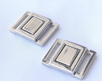 May include: Close-up of several silver-colored metal clasps and rings. Two square clasps are in focus, each with a rectangular center and a frame. Other clasps and rings are slightly blurred in the background, all on a white surface. These are likely jewelry findings.
