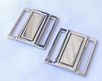 May include: Two silver metal bra strap adjusters, also known as bra extenders, on a white background. The adjusters are rectangular with a silver metal bar in the center.