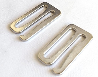 May include: A collection of silver-colored metal bra strap adjusters, scattered on a white surface. The adjusters have a rectangular shape with a curved hook at one end, designed for adjusting bra straps.