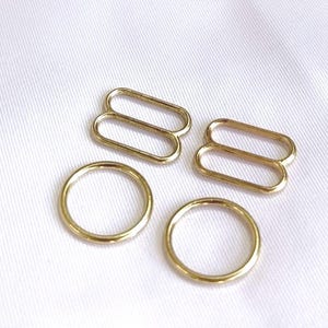 May include: Gold-coloured bra strap hardware, including rings and sliders, are scattered on a white fabric surface. The metal pieces are in various shapes and sizes, suitable for lingerie or garment construction.