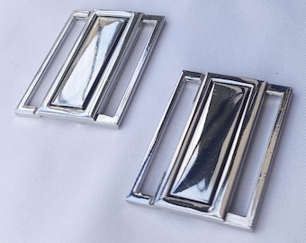 May include: Two silver metal slider buckles with a rectangular shape and a shiny finish. The buckles are designed for use in clothing or accessories.