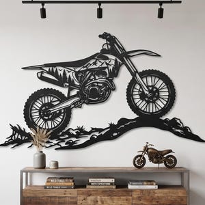 Può includere: Un'opera d'arte da parete in metallo nero che raffigura una moto da cross in movimento su un terreno accidentato. Il telaio della moto presenta un design ritagliato che raffigura un paesaggio montano. L'opera è montata su una parete liscia.