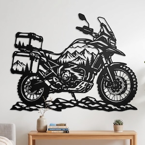 May include: A black metal wall art piece shaped like an adventure motorcycle. The motorcycle frame features a cutout design of a mountain range and pine trees. The bike includes side panniers, a front windshield, and spoked wheels.