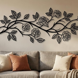 May include: Black metal wall art featuring a floral design of roses and leaves on a branch. The decorative piece is mounted on a beige wall above a gray sofa with pillows. The artwork adds a natural element to the room.