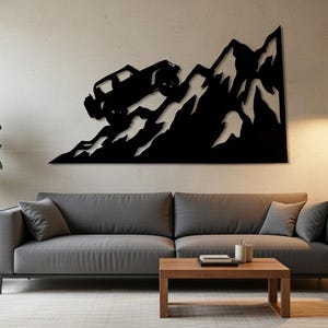 May include: Black metal wall art depicting a 4x4 vehicle climbing a mountain range. The artwork is mounted above a gray sofa in a modern living room setting. The wall is painted a neutral color.