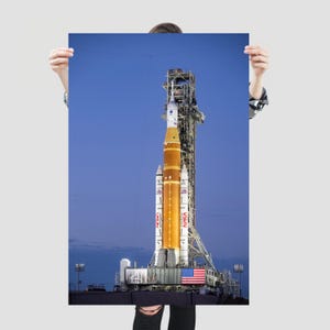 以下が含まれることがあります： 青い夕暮れの空を背景に、発射台に立つNASAのスペース・ローンチ・システム・ロケットが描かれた縦型のポスター。オレンジ色のコアステージの両脇には2基の白い固体燃料補助ロケットがあり、NASAのロゴとアメリカ国旗が確認できる。