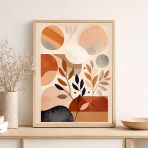 May include: Abstract art print in a light wood frame. The artwork features a beige background with geometric shapes in shades of orange, brown, and grey. Botanical elements are also included in the design. The print is displayed on a wooden surface.