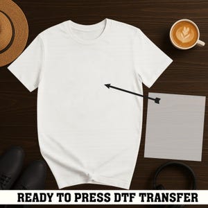 Pode incluir: Uma camiseta branca com gola redonda, pronta para uma transferência DTF. É exibida em uma superfície de madeira escura, junto com uma xícara de café, um chapéu de palha, sapatos pretos, fones de ouvido e uma folha de transferência cinza. Texto: READY TO PRESS DTF TRANSFER.