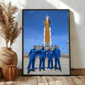以下が含まれることがあります： 青いフライトスーツを着た4人の宇宙飛行士が、澄んだ青空の下、発射台にある大きなオレンジと白のNASAロケットの前に立っている様子を描いた額入りアートプリント。プリントは細い黒いフレームに入っています。