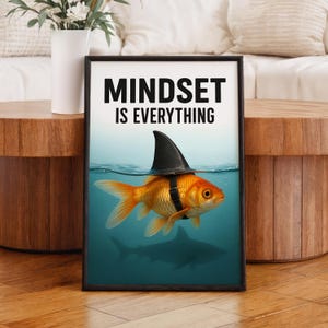 May include: Framed art print featuring a goldfish wearing a shark fin hat, swimming in blue water. The print has the text "MINDSET IS EVERYTHING" in bold black letters. The image is set against a wooden table.