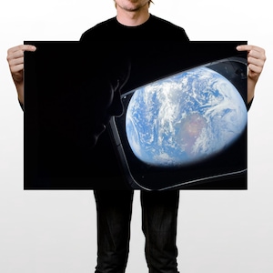 Peut inclure: Une impression murale rectangulaire présentant la silhouette sombre d'une personne regardant par une fenêtre rectangulaire arrondie vers une vue de la Terre depuis l'espace. La planète montre des océans bleus, des nuages blancs et des terres.