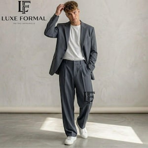 May include: A charcoal gray suit consisting of a single-breasted blazer with notched lapels and matching pleated trousers. The suit is worn over a plain white crew-neck t-shirt, paired with white sneakers. The Luxe Formal logo is visible.