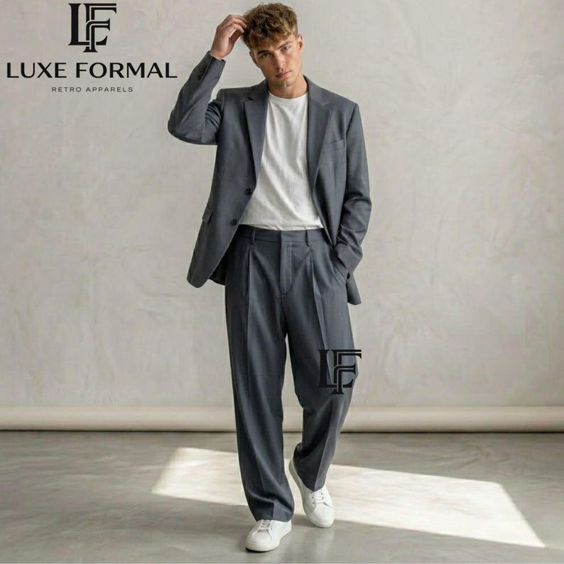 May include: A charcoal gray suit consisting of a single-breasted blazer with notched lapels and matching pleated trousers. The suit is worn over a plain white crew-neck t-shirt, paired with white sneakers. The Luxe Formal logo is visible.