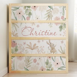 May include: A light wood bookshelf with three shelves. The panels are decorated with a floral pattern in pink, green, and beige. The name "Christine" is written in pink script. A cartoon bunny is also featured.