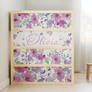 Può includere: Una libreria in legno con un design floreale con fiori viola e rosa, foglie verdi e il nome "Olivia" in corsivo. La libreria ha quattro sezioni ed è ambientata su uno sfondo bianco.