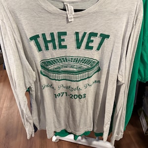 May include: A light grey long-sleeve t-shirt featuring a green graphic of a stadium. The text reads THE VET, Pride, Pretzels, Prison, and 1971-2003. The shirt has a crew neckline and is displayed on a black hanger against a rack of other clothing.