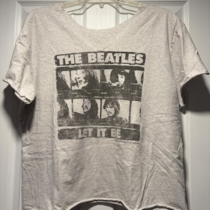 Könnte beinhalten: Hellgraues T-Shirt mit einem Schwarz-Weiß-Grafikdruck der Beatles. Der Text "THE BEATLES" steht darüber und "LET IT BE" darunter. Das Shirt hat einen rohen Saum und kurze Ärmel.
