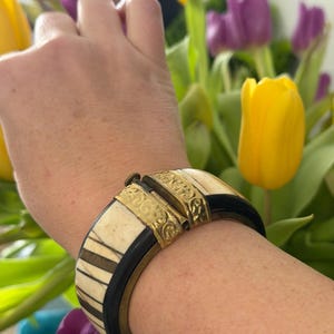 May include: A hinged bracelet with a cream, black, and gold design. The bracelet features cream-coloured rectangular segments, black bands, and gold-coloured metal accents. The bracelet is worn on a wrist, with a blurred floral background.