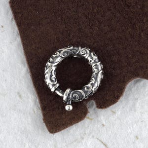 Könnte beinhalten: Ein silberfarbener, kreisförmiger Verschluss mit einem verzierten, wirbelnden Muster. Der Verschluss hat eine Scharnieröffnung und eine kleine, runde Kugel am Ende. Der Verschluss wird wahrscheinlich für Schmuck oder anderes Zubehör verwendet.