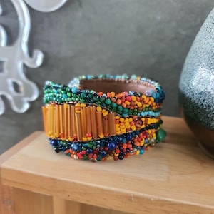 Puede incluir: Una pulsera de brazalete ancha con una densa disposición de cuentas de semillas multicolores en tonos verde, naranja, amarillo, azul y rojo. La sección central incluye una fila de cuentas tubulares naranjas translúcidas, con un forro de cuero beige.