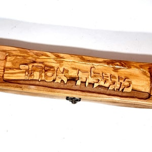 Op de afbeelding: Een houten doos met een scharnierend deksel, gesneden uit olijfhout. De doos heeft een donkerbruine afwerking en heeft Hebreeuwse letters op de bovenkant.