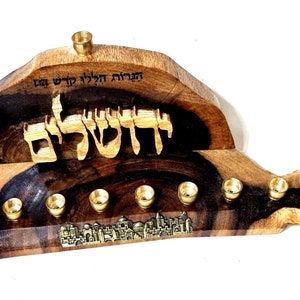 Op de afbeelding: Een houten menorah met een gesneden ontwerp van het Hebreeuwse woord "Jeruzalem" en een afbeelding van de skyline van de stad. De menorah heeft zeven kaarshouders en een kleine, aparte houder voor de sjamosj-kaars. De menorah is gemaakt van donker hout met een natuurlijke afwerking.