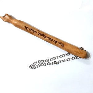 Może przedstawiać: Drewniany wskaźnik z ręcznie wykonanym wierzchołkiem i przymocowanym łańcuchem. Wskaźnik jest ozdobiony hebrajskim napisem "ברוך שם כבוד מלכותו לעולם ועד".