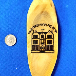 Puede incluir: Un letrero de madera de forma ovalada con una imagen en blanco y negro de una casa con el texto "770" y texto en hebreo. El letrero tiene un anillo plateado en la parte superior para colgar.