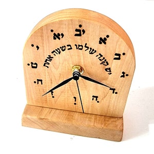 Op de afbeelding: Een houten klok met Hebreeuwse letters rond de wijzerplaat. De klok heeft zwarte wijzers en een gouden centrum. De klok staat op een houten voet.