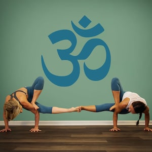 May include: Two people are performing a yoga pose, balancing on their hands with their legs intertwined. The pose is in front of a teal wall with a large blue Om symbol.