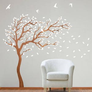 May include: A wall decal featuring a brown tree with white leaves and white birds flying around it. The tree is on a light gray wall with a white armchair in the foreground.