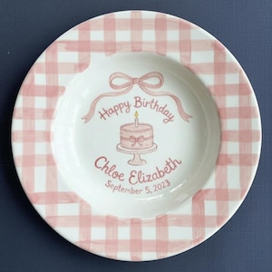 May include: A white ceramic plate with a pink gingham pattern around the rim. The centre has a pink birthday cake with a lit candle, a pink bow, and the words "Happy Birthday" and "Chloe Elizabeth September 5, 2023."