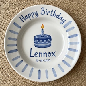 Könnte beinhalten: Weißer Keramikteller mit einer blauen Kuchenillustration und dem Text "Happy Birthday Lennox 12-18-2024". Der Teller hat blaue Pinselstrich-Akzente am Rand und eine brennende Kerze auf dem Kuchen.