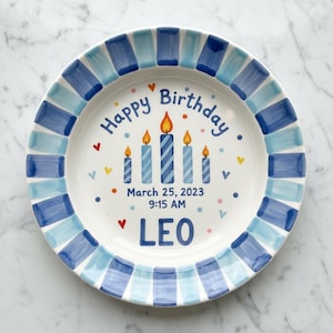 Pode incluir: Um prato de cerâmica branco com uma borda listrada azul e azul claro. O prato apresenta as palavras "Happy Birthday" e "Leo", juntamente com a data e a hora. Cinco velas são retratadas no centro, com pequenos corações e pontos espalhados.