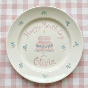 Pode incluir: Um prato de cerâmica branco com um design de bolo de aniversário rosa e azul. O prato tem as palavras "Happy Birthday" e o nome "Olivia" escrito. Pequenos detalhes florais azuis estão espalhados pela borda.