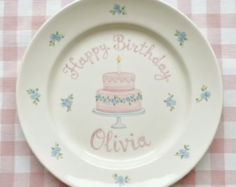 Puede incluir: Un plato de cerámica blanco con un diseño de tarta de cumpleaños rosa y azul. El plato tiene las palabras "Happy Birthday" y el nombre "Olivia" escrito. Pequeños detalles florales azules adornan el borde.