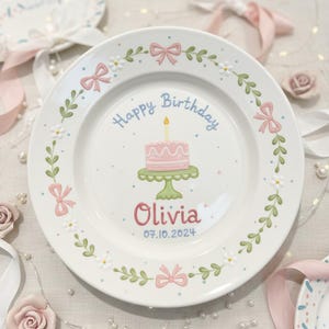 May include: A white ceramic plate with a pink cake illustration and the words "Happy Birthday" in blue. The name "Olivia" is in pink, with the date 07.10.2024 below. The plate is decorated with pink bows, green leaves, and white flowers.