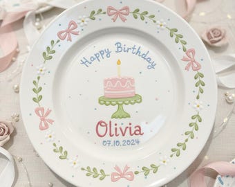 Puede incluir: Un plato de cerámica blanco con una ilustración de pastel rosa y las palabras "Happy Birthday" en azul. El nombre "Olivia" está en rosa, con la fecha 07.10.2024 debajo. El plato está decorado con lazos rosas, hojas verdes y flores blancas.
