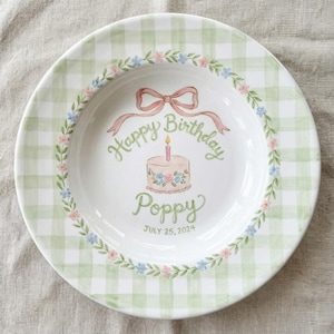 May include: A white ceramic plate with a green gingham pattern around the rim and floral details. The centre displays a birthday cake illustration with the text "Happy Birthday Poppy July 25, 2024". A pink bow is above the cake.