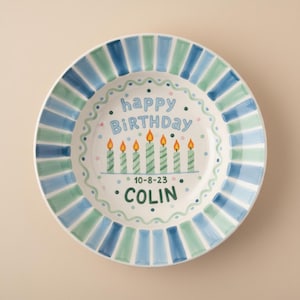 May include: A white ceramic plate with a blue and green striped border. The plate features the words "happy BIRTHDAY" and "COLIN" in blue lettering, along with six green candles with orange flames. The date "10-8-23" is also included.