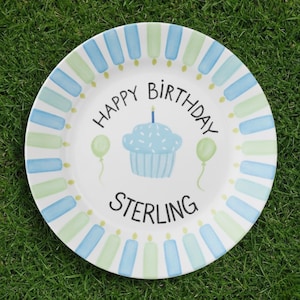 May include: White ceramic plate with a hand-painted birthday design. The plate features a blue cupcake with a candle, green and blue candles around the rim, and the words "Happy Birthday Sterling". Two green balloons are also painted on the plate.