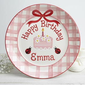 Puede incluir: Un plato redondo de cerámica blanca con un borde a cuadros rojos. El centro presenta un pastel rosa con margaritas, una vela encendida y las palabras "Happy Birthday Emma" en rojo. Dos mariquitas y un lazo rojo completan el diseño.