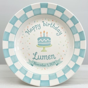 May include: A white ceramic plate with a blue and white chequered border. The centre features a blue cake with three candles, the words "Happy birthday" and "Lumen November 1, 2021". The background is decorated with colourful confetti.