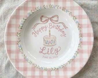 May include: A white ceramic plate with a pink gingham border. The center features a cake with a lit candle, the words "Happy Birthday Lily" and the date "June 10, 2025". A floral garland encircles the inner rim.