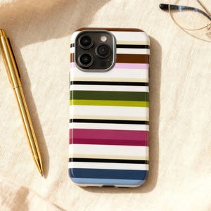 May include: A smartphone case with a colorful striped pattern in white, brown, pink, green, black, and blue. A gold pen and eyeglasses are also visible on a beige surface.