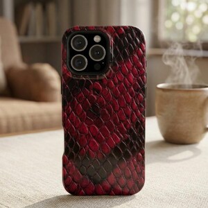 Puede incluir: Una funda para smartphone con un patrón de piel de serpiente en rojo oscuro y negro. La funda tiene un recorte para un sistema de cámara de triple lente en la parte posterior. La superficie texturizada imita la apariencia de escamas.
