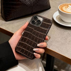 Könnte beinhalten: Eine braune Handtasche mit Krokodiltextur und eine passende Handyhülle. Die Handtasche hat ein geschwungenes Design und einen einzelnen Riemen. Eine Tasse Kaffee mit Latte Art steht auf einem Tisch.