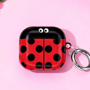 May include: A red and black ladybug-themed AirPod case with a silver ring, surrounded by colorful beaded bracelets, stickers, and a retro camera on a pink background.